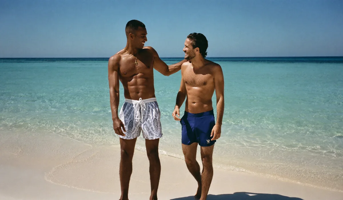 Twee lachende mannen op het strand, de ene in een ruime zwemshort, de andere in een strakke zwemshortort